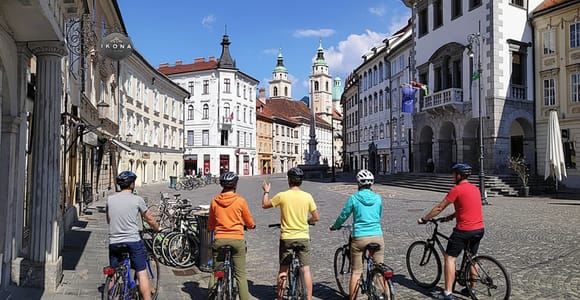 Five cyclists in helmets stand with their bikes on a cobbled square, facing pastel old town houses and church towers under a blue sky