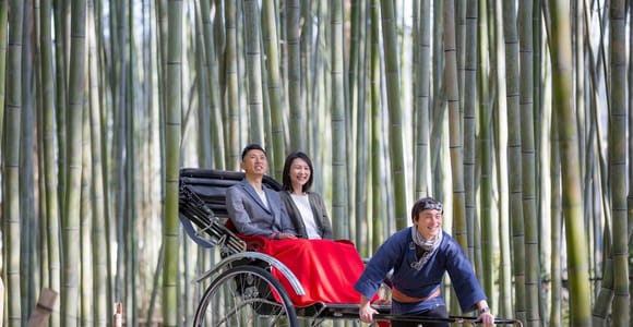 Two passengers seated under a red blanket in a black rickshaw, the puller in a blue happi coat gripping the shafts, tall bamboo behind