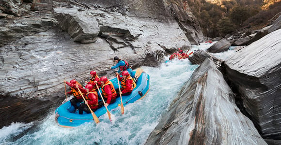 A raft crew in helmets and red life jackets paddling through white water between rocky canyon walls