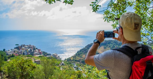 Hiker with a red backpack photographing a pastel village on a headland far below, with the sea glittering behind