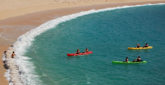 Three kayaks on turquoise water in a curved bay, a swimmer standing in the white shorebreak on the empty sand