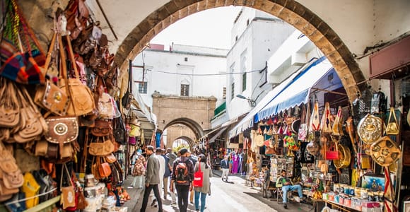 A medina lane under a stone arch lined with leather bags and craft stalls, people walking between whitewashed walls