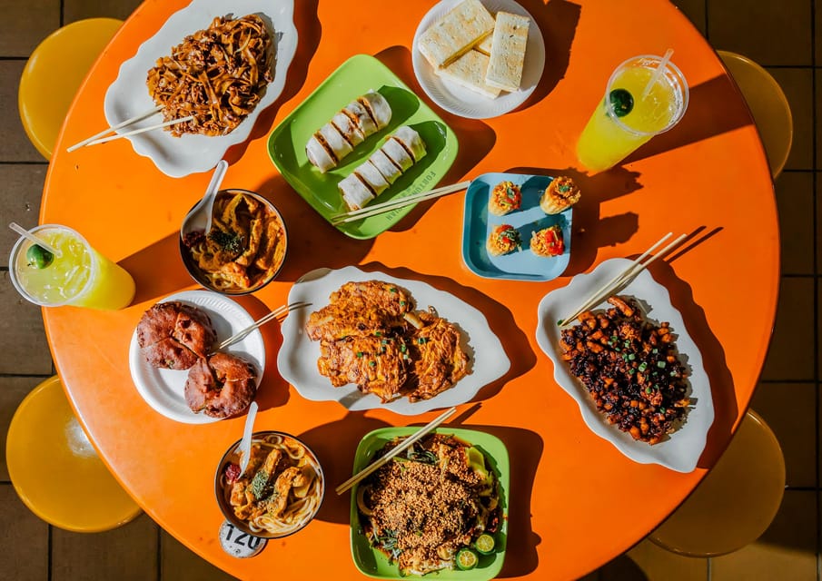 Overhead view of an orange table covered with plates of noodles, dumplings, satay and two citrus drinks