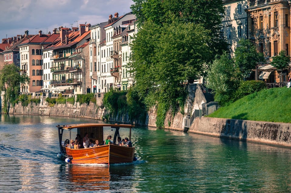 A wooden boat with passengers glides along a green river past old town houses and a tree-lined stone embankment