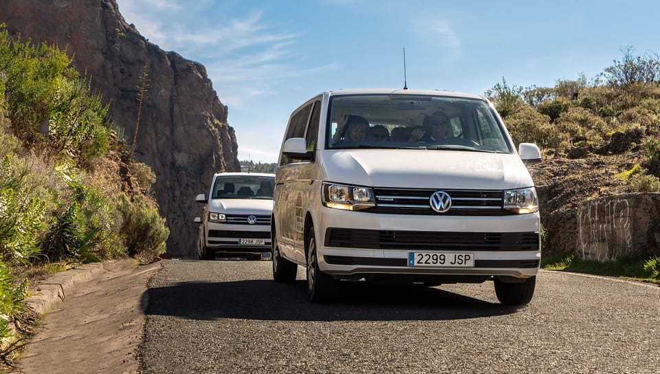 Two white minibuses coming down a narrow tarmac road cut into a rock face