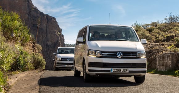 Two white minibuses coming down a narrow tarmac road cut into a rock face