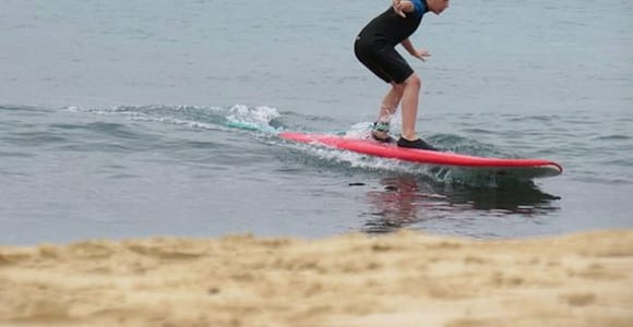 A child in a wetsuit standing up on a red foam board in small shore waves, moored boats behind
