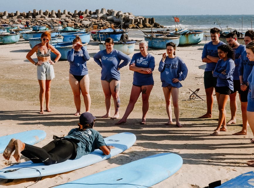 A surf instructor lying on a board on the sand demonstrating to a semicircle of students, round woven fishing boats pulled up behind them