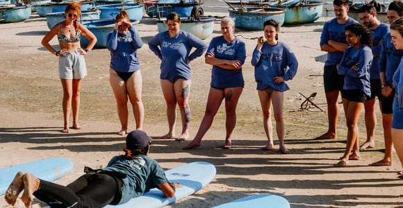A surf instructor lying on a board on the sand demonstrating to a semicircle of students, round woven fishing boats pulled up behind them
