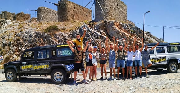 Two black and white Land Rover safari vehicles parked at a rocky hilltop with old stone windmills, a group of people with raised arms in front