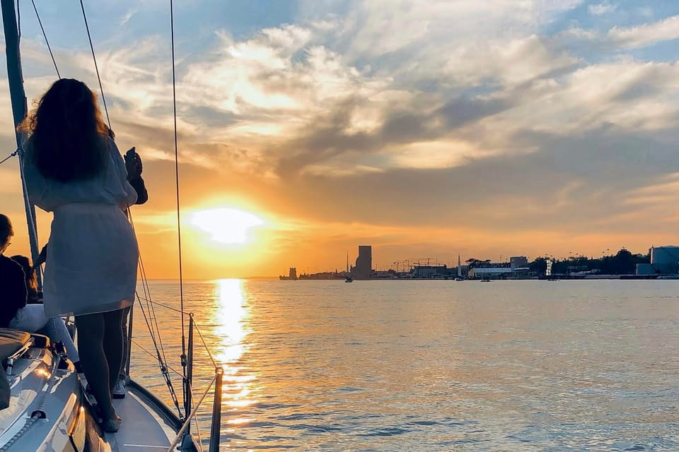 The foredeck of a sailing yacht on the Tagus at sunset, a passenger standing at the rail taking a photograph, the silhouette of the Torre de Belém on the horizon