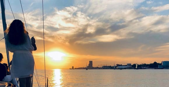 The foredeck of a sailing yacht on the Tagus at sunset, a passenger standing at the rail taking a photograph, the silhouette of the Torre de Belém on the horizon