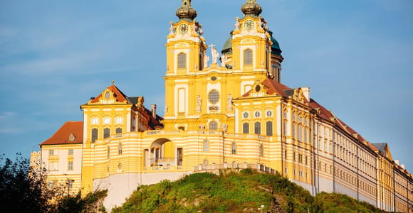 The yellow and white baroque Melk Abbey with twin towers on a wooded hill under a blue sky