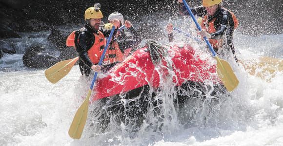 A red raft with four helmeted paddlers hitting a wave of white water