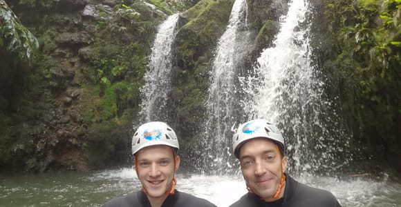 Two people in helmets and wetsuits standing in a pool below a forest waterfall
