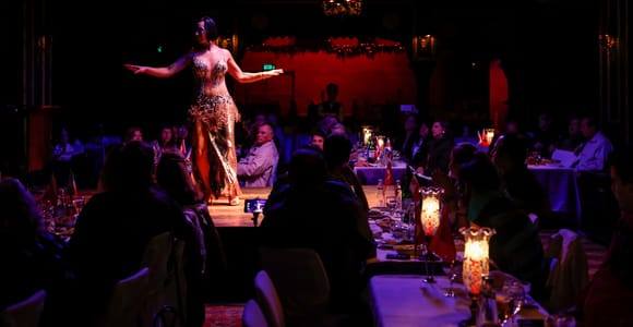 A dancer in a sequinned costume performs on a stage between laid tables in a darkened restaurant