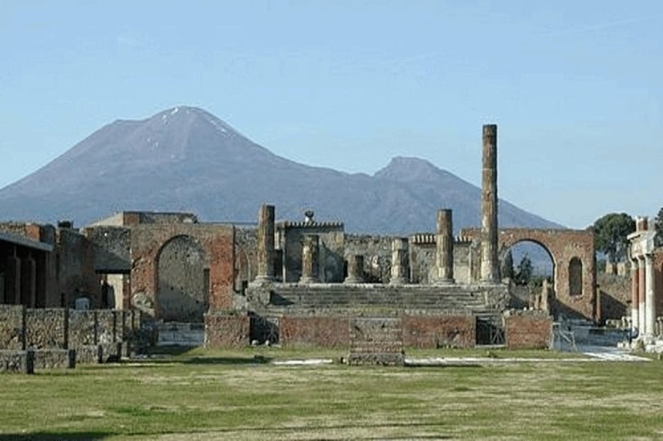 Ruins with columns and arches on a grassy field, with Mount Vesuvius rising behind them