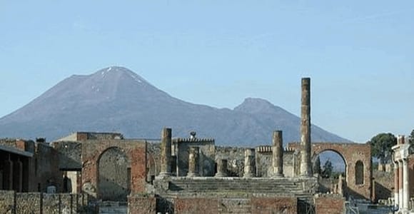 Ruins with columns and arches on a grassy field, with Mount Vesuvius rising behind them