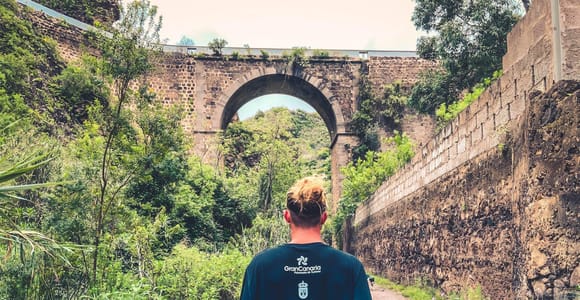 A walker approaching a stone bridge amid dense green vegetation