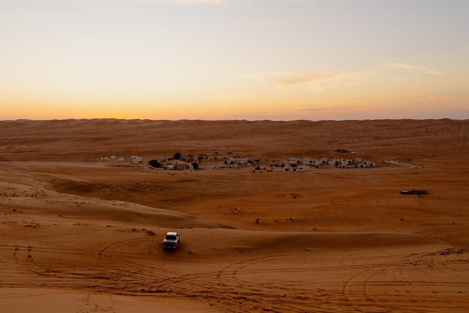 Dusk over the Wahiba dunes: a white 4x4 on a dune crest in the foreground casting tyre tracks across the orange sand, a scattering of low white camp buildings in the hollow below, and a pale gold horizon