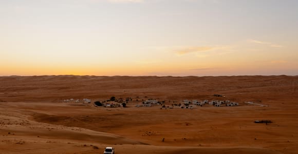 Dusk over the Wahiba dunes: a white 4x4 on a dune crest in the foreground casting tyre tracks across the orange sand, a scattering of low white camp buildings in the hollow below, and a pale gold horizon