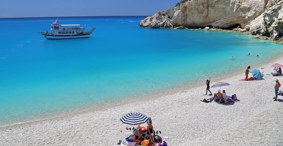 An excursion boat anchored in turquoise water off a white pebble beach with sunshades, beneath a pale limestone headland