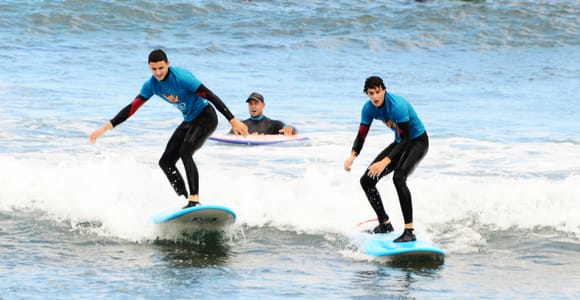 Beginners carrying foam surfboards across a sandy beach towards the sea