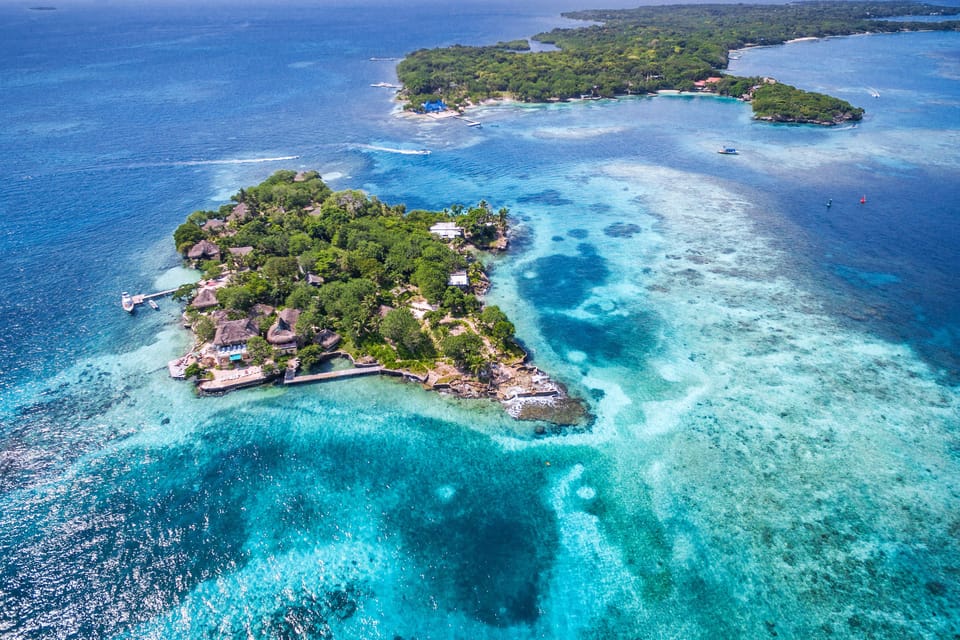Aerial view of a small wooded island with thatched roofs and a jetty, ringed by pale turquoise shallows and dark patches of reef, a larger low island behind it
