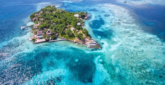 Aerial view of a small wooded island with thatched roofs and a jetty, ringed by pale turquoise shallows and dark patches of reef, a larger low island behind it