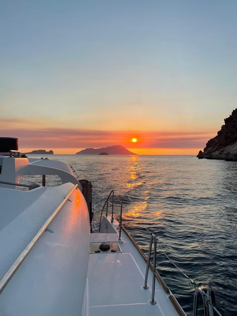 Orange sunset over calm sea seen from the deck of a boat, with a rocky headland on the right and islands on the horizon