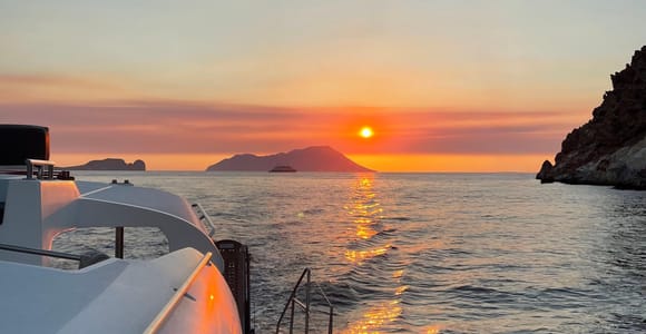 Orange sunset over calm sea seen from the deck of a boat, with a rocky headland on the right and islands on the horizon