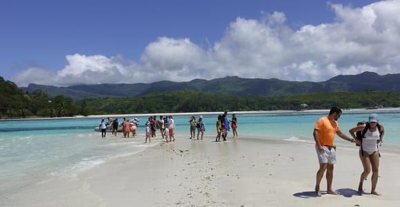 People wading onto a bright sandbank in shallow turquoise water, with green mountains and towering clouds behind