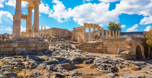 Ancient stone columns and a ruined portico on the Lindos acropolis, with rocky ground in front and a blue sky full of white clouds above