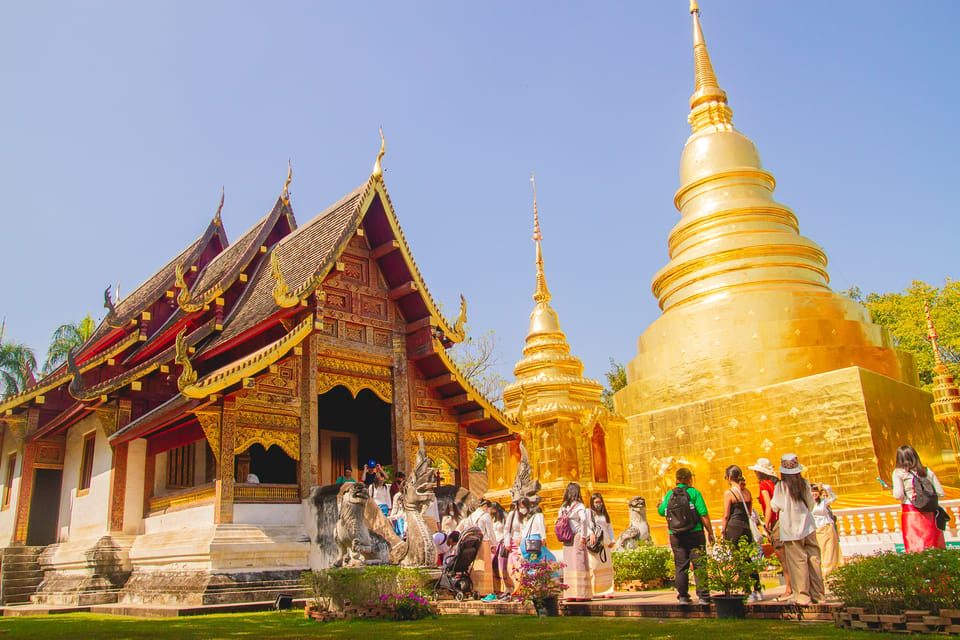 A gilded bell-shaped chedi and a red-and-gold Lanna hall side by side, visitors gathered on the paving between them under a pale sky