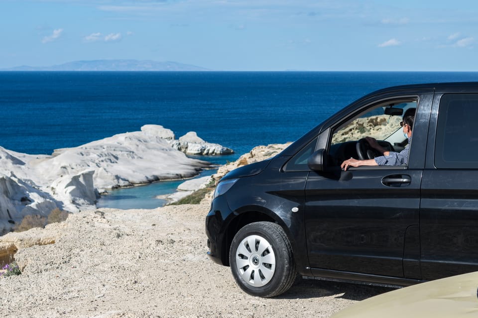 A black minivan with its driver at a cliff edge above the white rock coast of Sarakiniko