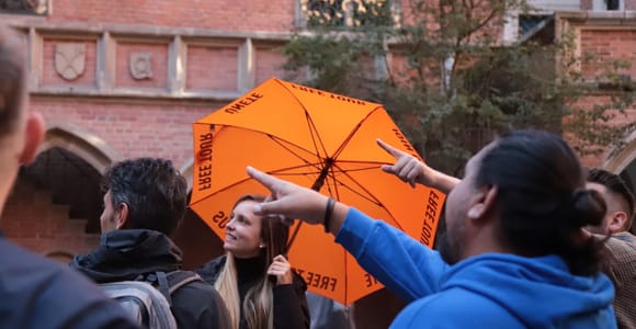 A man in a blue hooded top points ahead beside a woman holding an orange umbrella printed with the words free tour, in a brick courtyard