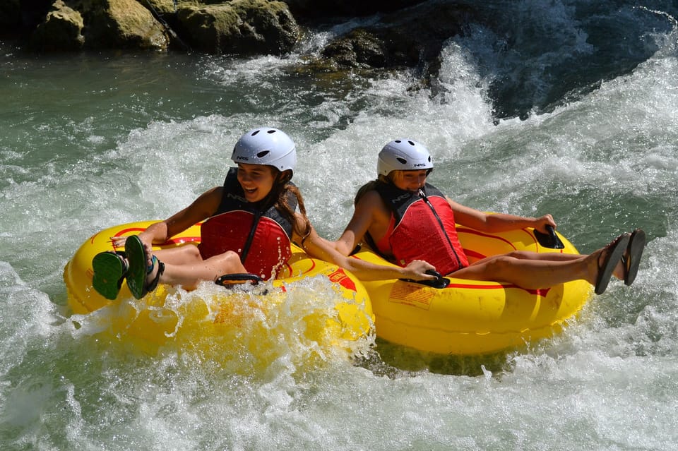 Two young people in helmets and red life jackets riding yellow inflatable tubes through white water, laughing