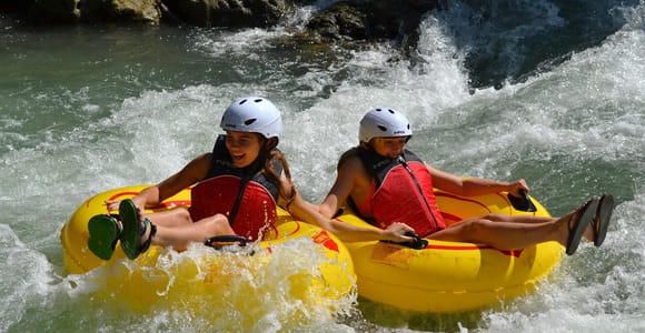 Two young people in helmets and red life jackets riding yellow inflatable tubes through white water, laughing