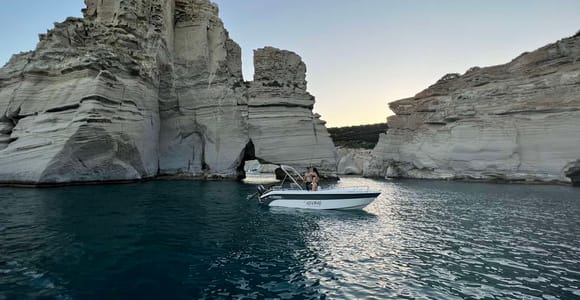 Small motorboat anchored in clear turquoise water beside a pale rock cliff, with swimmers near the stern
