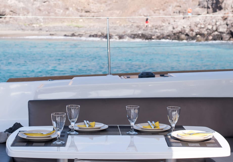 Tisch mit gedecktem Essen und Getränken auf einem Katamaran, mit Blick auf das Meer und die Küste von Fuerteventura im Hintergrund.