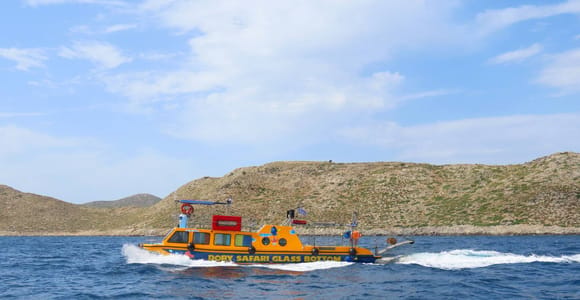 A small yellow and blue boat with a glass-bottom sign speeding past a bare brown island under a blue sky