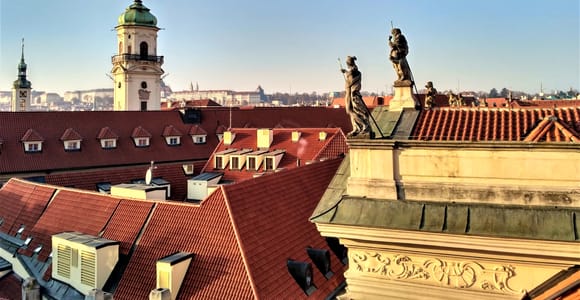 Red tiled rooftops of Prague Old Town with baroque statues in the foreground and a white tower with a green dome behind