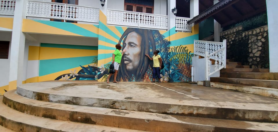 A large painted portrait of Bob Marley on a wall of yellow and turquoise sunburst rays, two visitors standing in front of it on a curved concrete terrace