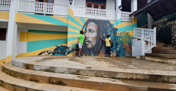 A large painted portrait of Bob Marley on a wall of yellow and turquoise sunburst rays, two visitors standing in front of it on a curved concrete terrace
