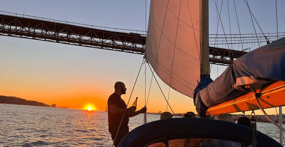 A man standing at the bow of a small sailing yacht holding a bottle, the sail filled above him, the sun setting beneath the girders of a road bridge