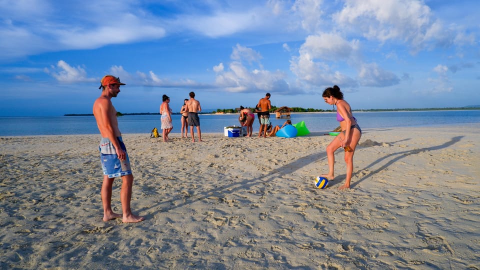 People standing on a wide strip of white sand in late afternoon light, one woman about to kick a volleyball, shallow blue sea and tall cumulus clouds behind