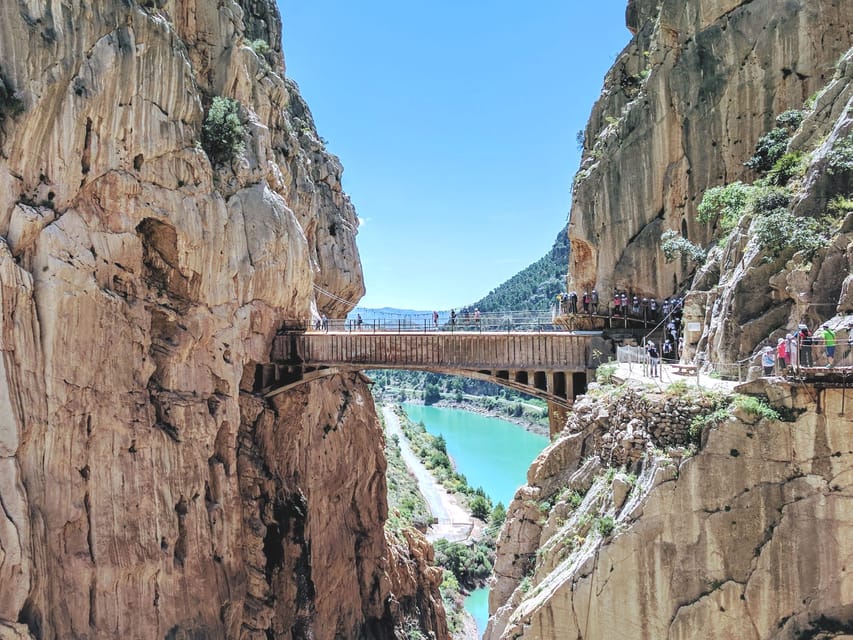 A footbridge across a deep rocky gorge, with walkers on the cliffside path and turquoise water far below