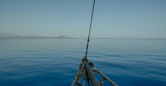 The bow of a sailing ship with a rope net above calm blue sea and a distant coastline on the horizon