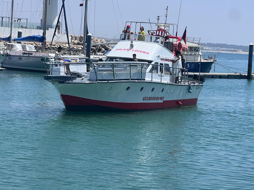 A white excursion boat with a red hull stripe moored in a calm marina, a handful of passengers on the upper deck, sailing yachts and a breakwater behind
