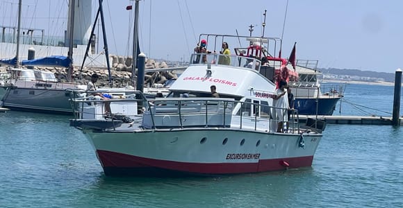 A white excursion boat with a red hull stripe moored in a calm marina, a handful of passengers on the upper deck, sailing yachts and a breakwater behind